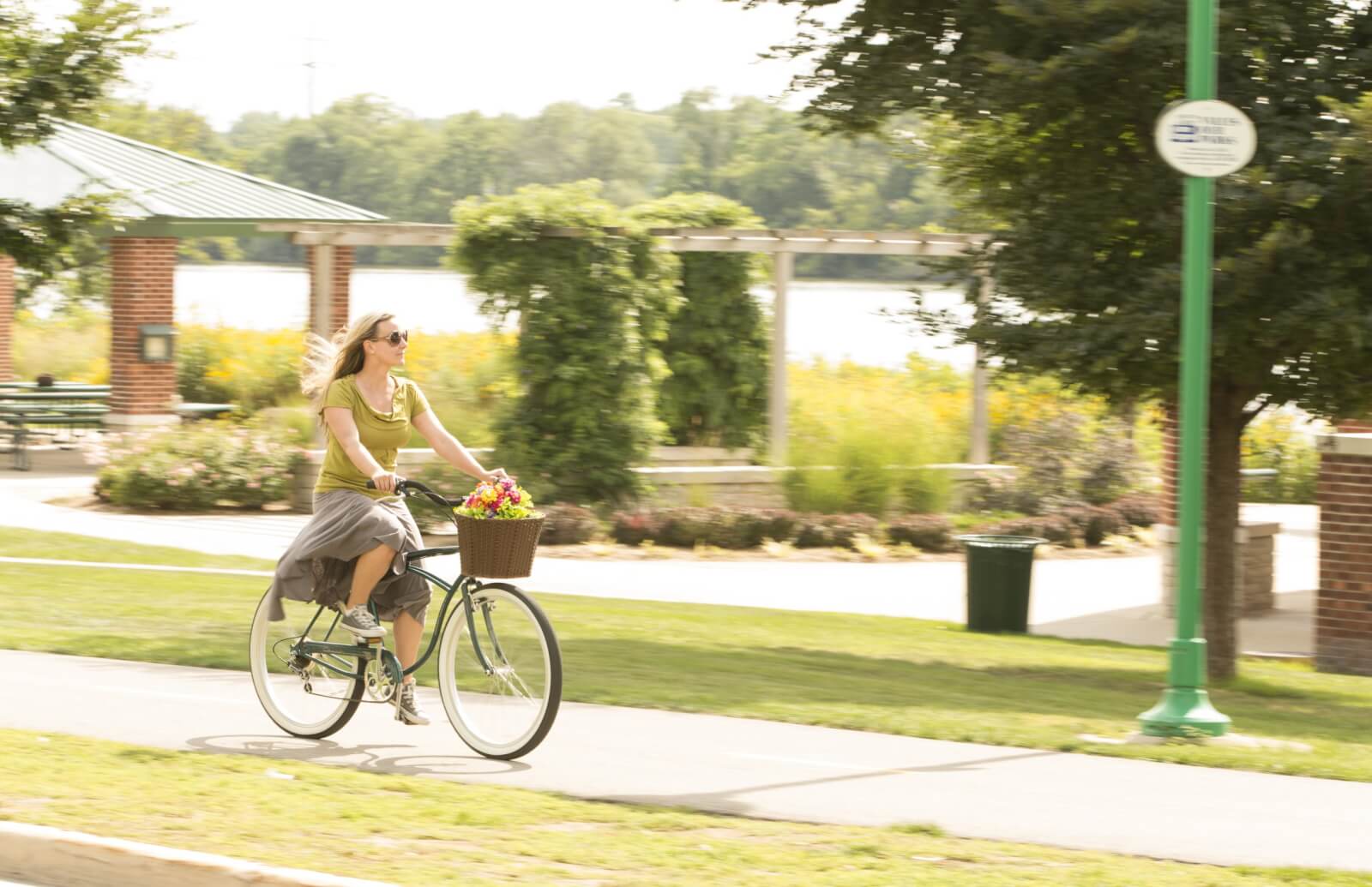 Things to Do A woman rides a bike with a basket full of flowers along a bike path in a park on a sunny day.