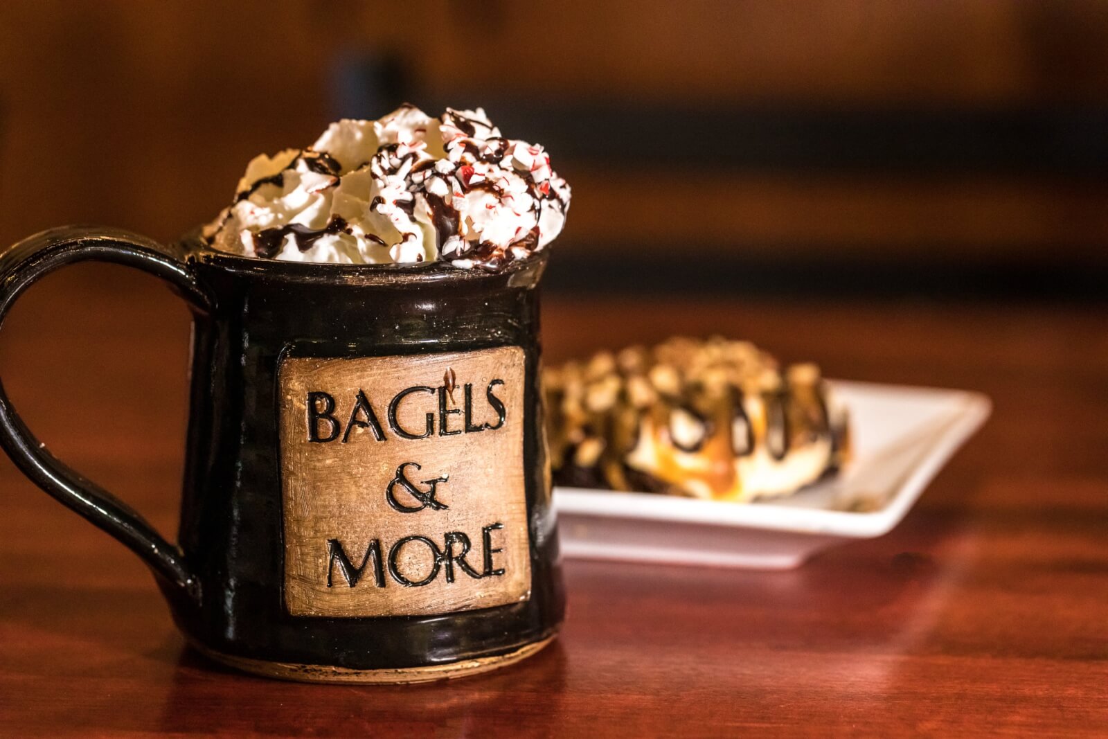 Plan Your Trip Bagels and More coffee cup is topped with whipped cream with chocolate drizzle and peppermint crumbs sits in front of a plate of pastry.